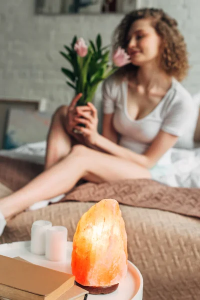 Selective focus with young woman holding flowers in bedroom with Himalayan salt lamp — Stock Photo