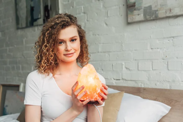 Happy curly girl in homewear holding Himalayan salt lamp in bedroom — Stock Photo