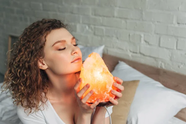 Tender woman with closed eyes in homewear holding Himalayan salt lamp in bedroom — Stock Photo