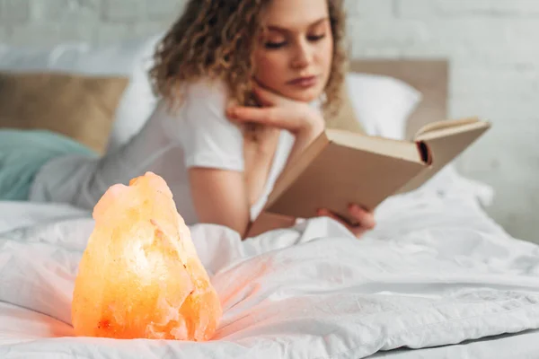 Foco seletivo da mulher encaracolada ler livro na cama com lâmpada de sal do Himalaia — Fotografia de Stock