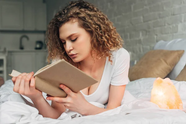 Attraente libro di lettura ragazza riccia sul letto con lampada al sale himalayano — Foto stock