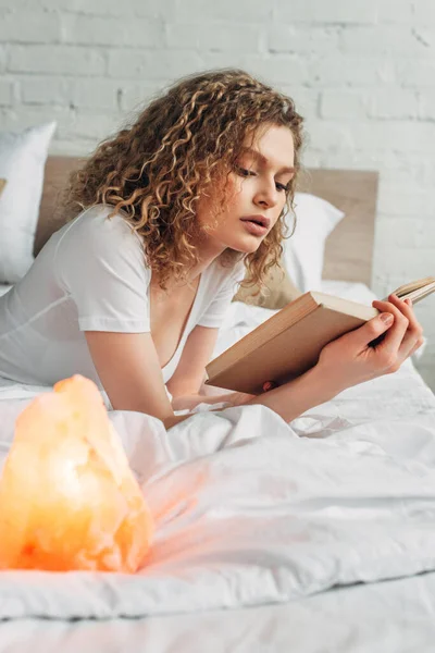 Chica rizada leyendo libro en la cama con lámpara de sal del Himalaya, enfoque selectivo - foto de stock