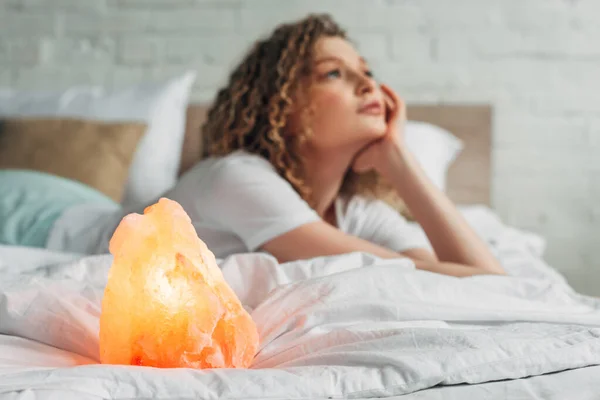 Fille réfléchie couchée sur le lit avec la lampe de sel himalayenne, foyer sélectif — Photo de stock