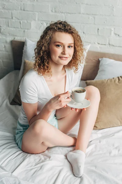 Sonriente chica rizada en ropa de casa sosteniendo taza de café en la cama por la mañana - foto de stock