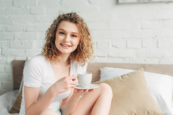 Привлекательная счастливая кудрявая девушка в домашней одежде держит чашку кофе на кровати утром — стоковое фото