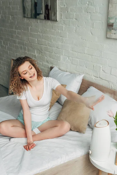 Cheerful beautiful girl on bed with air purifier spreading steam — Stock Photo