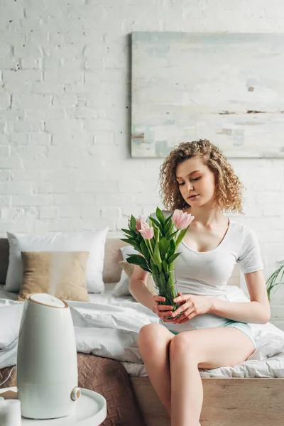 Приваблива жінка тримає квіти, сидячи в спальні з очищувачем повітря — стокове фото
