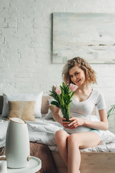 Ragazza felice che tiene i tulipani mentre si siede in camera da letto con purificatore d'aria — Foto stock