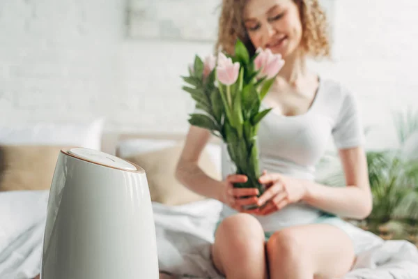 Вибірковий фокус усміхненої дівчини, що тримає квіти тюльпанів, сидячи в спальні з очищувачем повітря — стокове фото