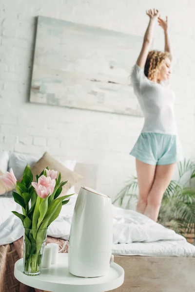 Mujer estiramiento en dormitorio con purificador de aire y flores, enfoque selectivo - foto de stock