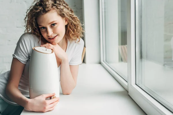 Hermosa chica feliz con purificador de aire en alféizar de la ventana - foto de stock