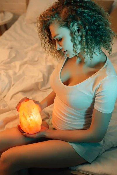 Кудрявая девушка с гималайской соляной лампой, сидя в постели вечером — стоковое фото