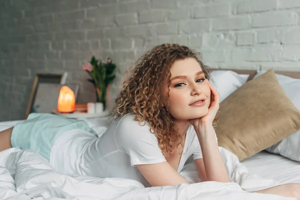 Smiling girl in homewear lying on bed in bedroom with Himalayan salt lamp — Stock Photo