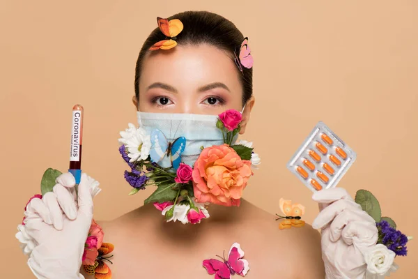 Nua menina asiática em luvas de látex e máscara facial floral com borboletas segurando pílulas e coronavírus teste de sangue isolado no bege — Fotografia de Stock