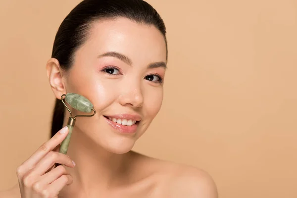 Attractive naked asian woman making massage of face with jade roller isolated on beige — Stock Photo