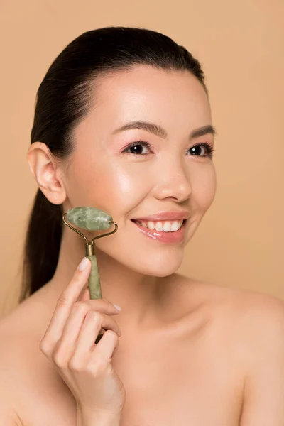 Happy naked asian girl making massage of face with jade roller isolated on beige — Stock Photo