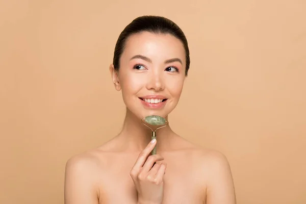 Hermosa alegre asiático chica haciendo masaje de cara con jade rodillo aislado en beige - foto de stock