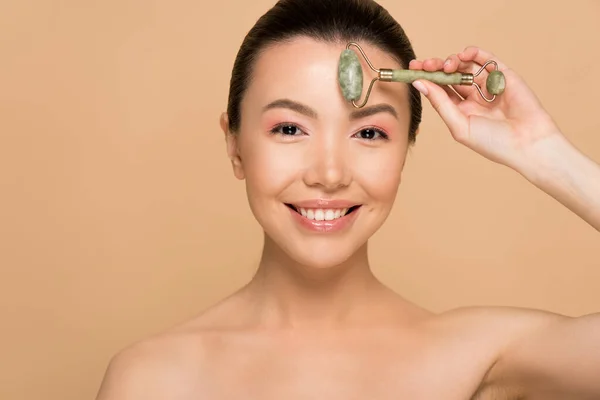 Beautiful nude asian woman making massage of face with jade roller isolated on beige — Stock Photo