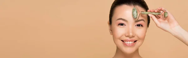 Plan panoramique de belle fille asiatique souriante faisant massage du visage avec rouleau de jade isolé sur beige — Photo de stock