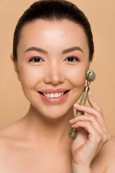Beautiful smiling naked asian girl making massage of face with jade roller isolated on beige — Stock Photo