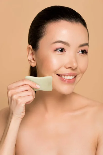 Hermosa feliz desnudo asiático mujer haciendo masaje de cara con jade gua sha piedra aislado en beige - foto de stock