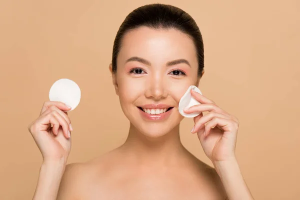 Smiling naked asian girl removing makeup from face with cotton pads isolated on beige — Stock Photo