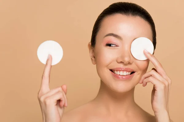 Happy naked asian girl removing makeup from face with cotton pads isolated on beige — Stock Photo