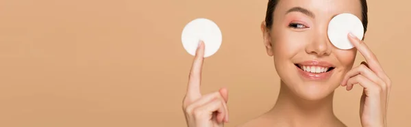 Plano panorámico de sonriente chica asiática quitando el maquillaje de la cara con almohadillas de algodón aislado en beige - foto de stock