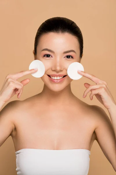 Cheerful asian girl removing makeup from face with cotton pads on beige — Stock Photo