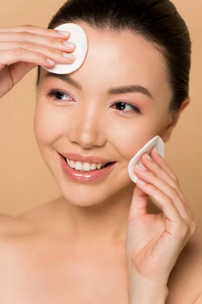 Attractive smiling nude asian woman removing makeup from face with cotton pads isolated on beige — Stock Photo