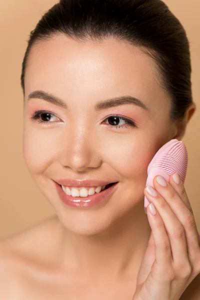 Beautiful smiling naked asian girl using silicone cleansing facial brush isolated on beige — Stock Photo