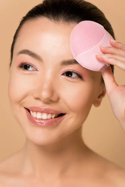 Attractive smiling asian girl using pink silicone cleansing facial brush isolated on beige — Stock Photo