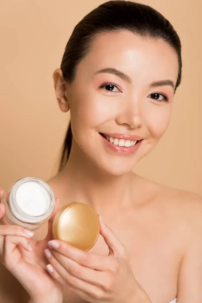 Beautiful smiling naked asian girl holding glass container with cosmetic cream isolated on beige — Stock Photo