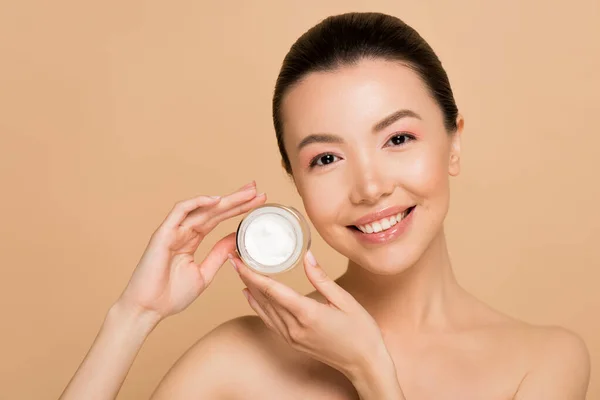 Smiling naked asian woman holding glass container with cosmetic cream isolated on beige — Stock Photo