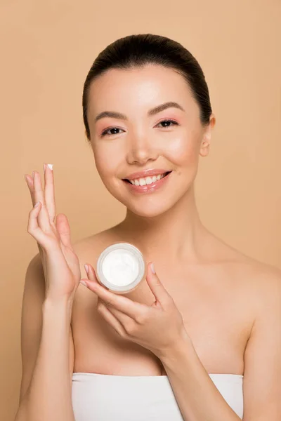 Attraente sorridente asiatico ragazza holding vetro contenitore con crema idratante isolato su beige — Foto stock