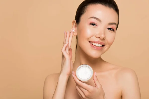 Beautiful nude asian woman applying cosmetic cream on face isolated on beige — Stock Photo