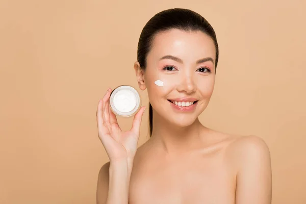 Hermosa sonriente chica asiática desnuda sosteniendo contenedor de vidrio con crema cosmética aislado en beige - foto de stock