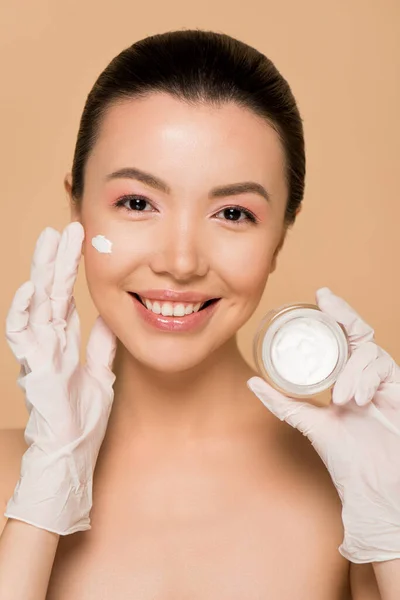 Beautiful happy nude asian girl in latex gloves applying face cream isolated on beige — Stock Photo