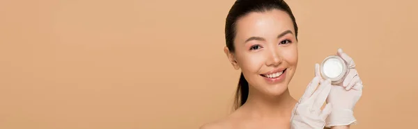 Panoramic shot of smiling nude asian girl in latex gloves holding glass container with face cream isolated on beige — Stock Photo