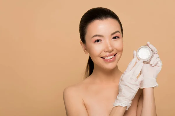 Attractive happy nude asian girl in latex gloves holding glass container with face cream isolated on beige — Stock Photo