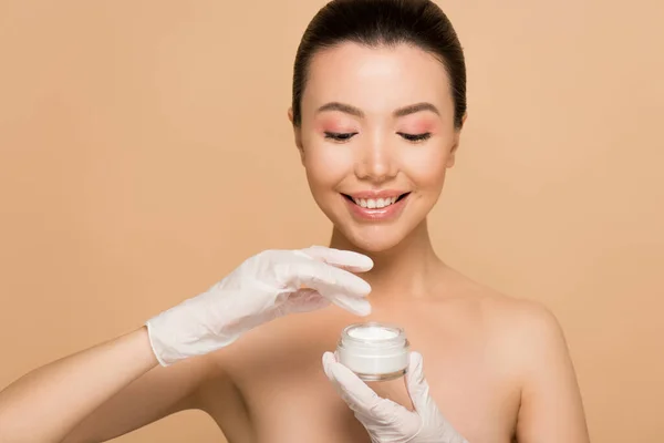 Attractive happy nude asian girl in latex gloves applying face cream isolated on beige — Stock Photo