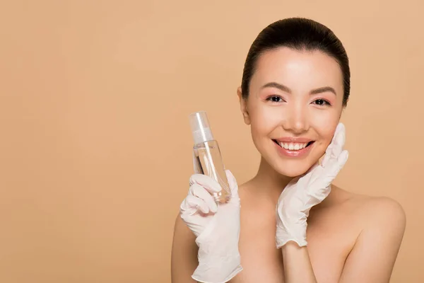 Attractive cheerful nude asian girl in latex gloves holding antiseptic spray isolated on beige — Stock Photo