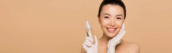 Panoramaaufnahme einer fröhlichen nackten asiatischen Frau in Latex-Handschuhen, die antiseptisches Spray isoliert auf beige hält — Stockfoto