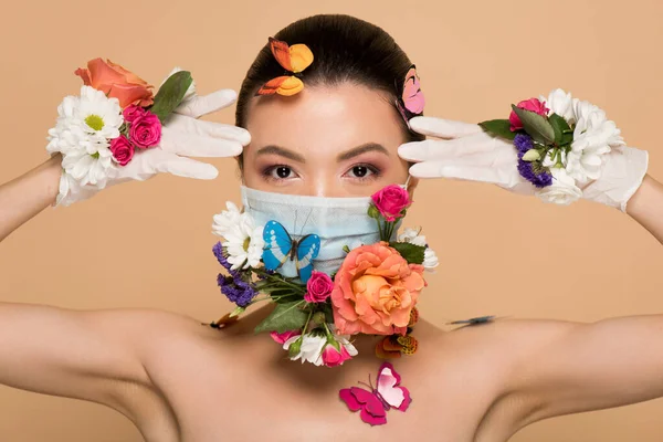 Atractiva mujer asiática en guantes de látex y mascarilla floral con mariposas aisladas en beige - foto de stock