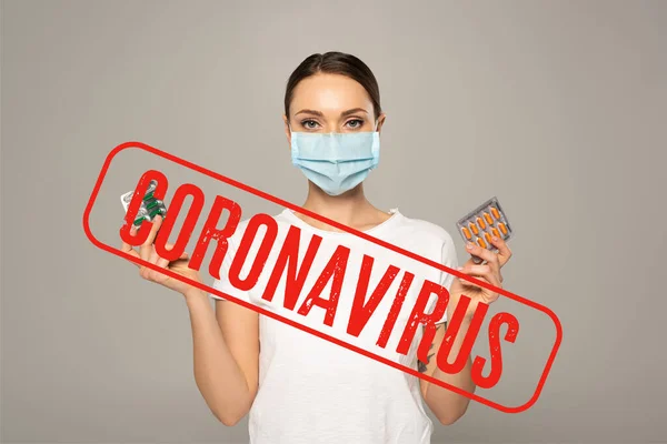 Женщина в медицинской маске держит волдыри с таблетками, изолированными на серой, коронавирусной иллюстрации — стоковое фото