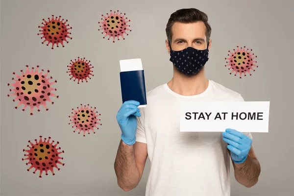 Hombre con máscara de seguridad y guantes de látex que sostiene la tarjeta con letras de estancia en casa y pasaporte con billete de avión aislado en gris, ilustración de bacterias - foto de stock