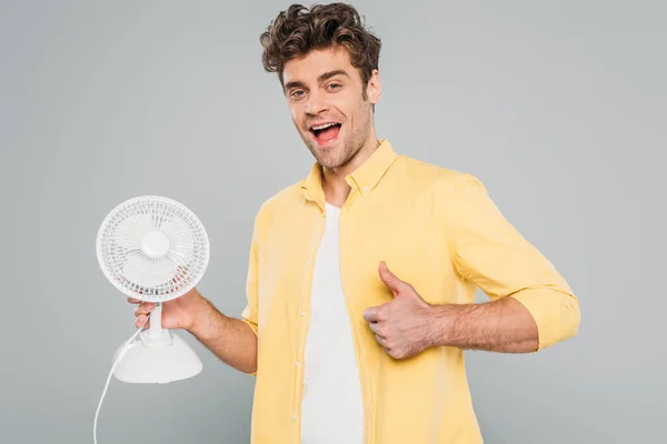 Захоплений чоловік з настільним вентилятором дивиться на камеру і показує як знак ізольований на сірому — стокове фото