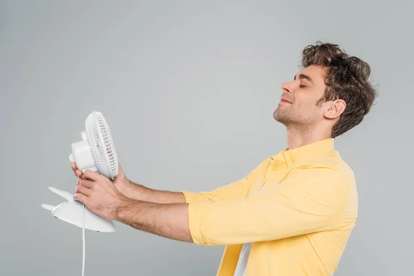 Uomo con gli occhi chiusi e le mani tese tenendo ventilatore scrivania e sorridente isolato su grigio — Foto stock