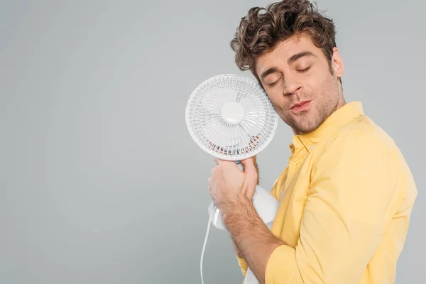 Hombre con los ojos cerrados sosteniendo ventilador de escritorio aislado en gris - foto de stock