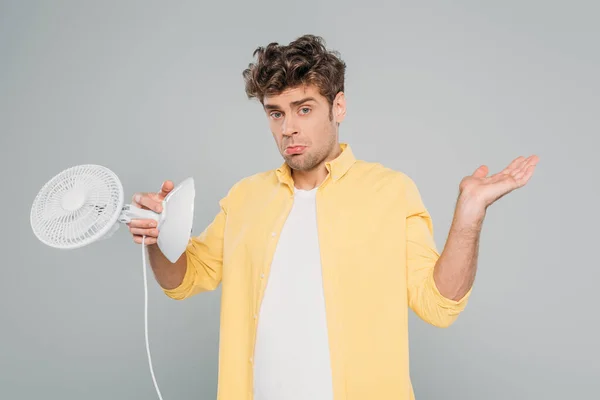 Vista frontale dell'uomo confuso che tiene il ventilatore della scrivania e guarda la telecamera isolata sul grigio — Foto stock